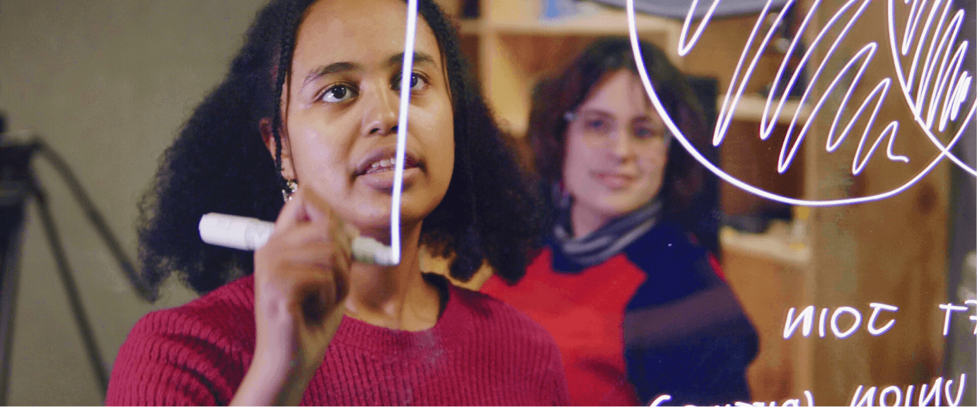 A headshot of a Black, female engineer drawing a diagram on a transparent whiteboard, viewed through the board itself