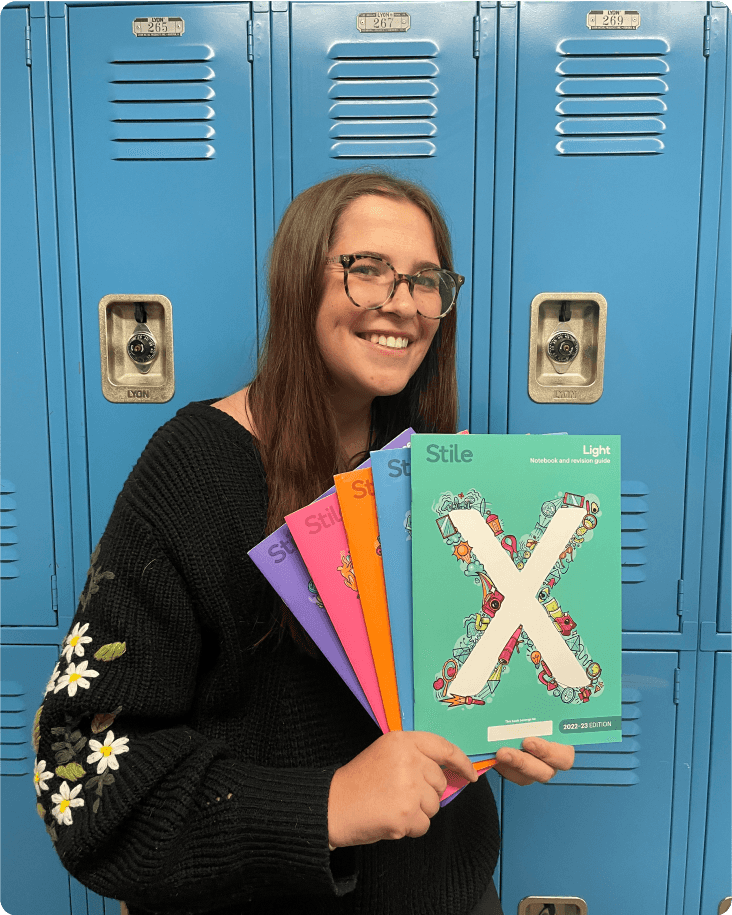 A teacher holds up a stack of Stile X booklets, smiling at the camera. She is standing in front of blue school lockers