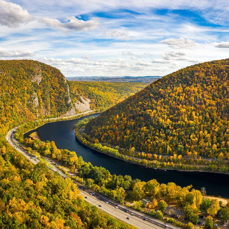 A wide aerial view of a river winding between two forested hills covered in autumn foliage, with a highway running alongside the water.