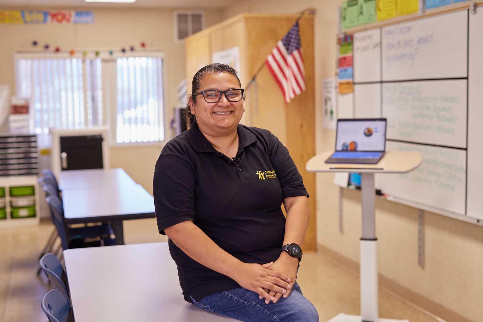 Jovana is shown sitting on a table in her classroom, smiling at the camera