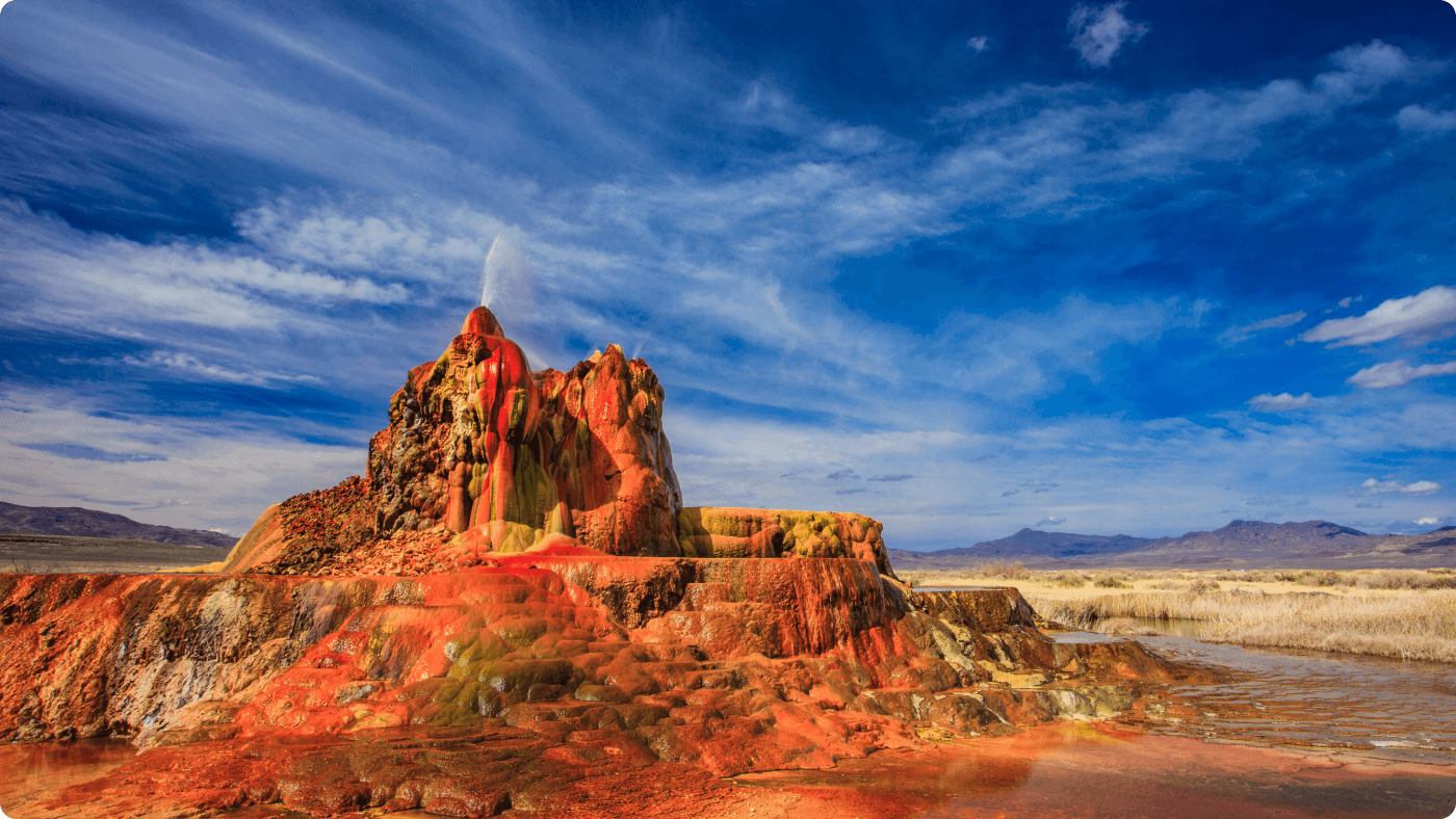 A large, red rock outcropping in the foreground, with a spout of water coming out of the top. The background is bright blue sky and open plains