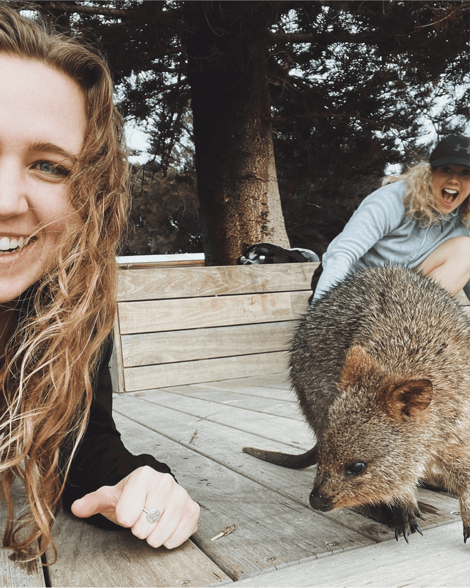 A selfie including half the photographer's face. She is up close with a quokka, and another team member looks excited in the background