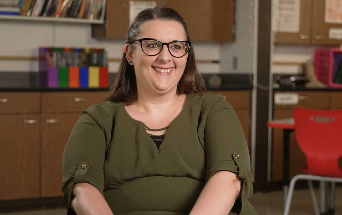 Person with glasses smiling while seated in a classroom, with shelves and colorful boxes in the background.